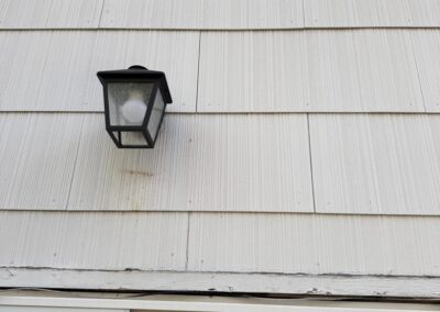 A black outdoor wall lantern is mounted on white siding above a window with closed blinds.