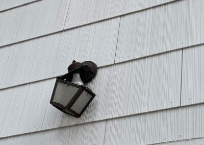 A black outdoor wall lantern with a white bulb is mounted on light-colored siding. The lantern is tilted slightly downward.